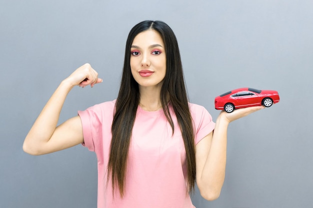 Driving school idea and concept, student driver passed the\
exam, drivers license, portrait of a beautiful happy young woman,\
holding a car in her hand. on a gray isolated background. shows his\
muscles