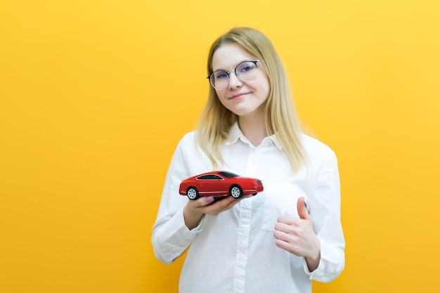 自動車教習所のアイデアとコンセプトの学生の運転手は、車を見て黄色の背景に彼女の手で車を持っている美しい幸せな若い女性の試験運転免許証の肖像画に合格しました