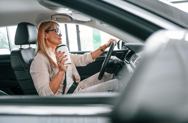 Driving vehicle and holding cup of drink Woman in white formal clothes is in the car dealership