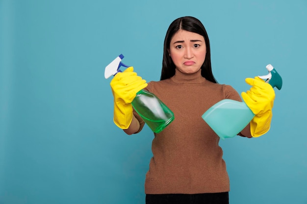 Droevig boze jonge aziatische vrouw die twee wasmiddelnevel in gele handschoenen houdt die op een blauwe achtergrond worden geïsoleerd, huisconcept schoonmaken, ongelukkig depressief en uitgeputte negatieve huishoudster