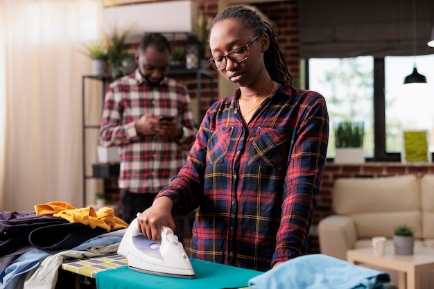 Droevige afro-amerikaanse vrouw die thuis kleding strijkt, huisvrouw die voor het huishouden zorgt terwijl de afgeleide echtgenoot mobiele telefoon gebruikt. Vermoeide drukke gefrustreerde ontevreden vrouw met alle huishoudelijke klusjes