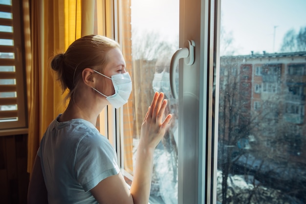 Droevige eenzame vrouw in beschermend medisch masker op haar gezicht dat venster bekijkt