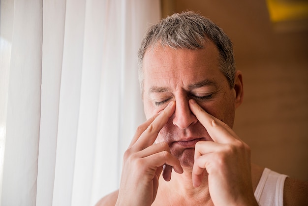 Droevige en depressieve man houdt zijn hoofd met zijn hand, staand bij het raam. De man houdt zijn neus- en sinusgebied met vingers in duidelijke pijn van een hoofdpijn in het voorhoofdgebied