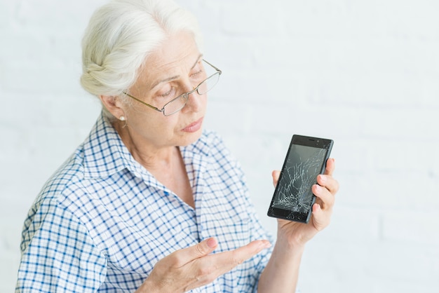 Droevige hogere vrouw die smartphone met het gebroken scherm tonen tegen witte achtergrond