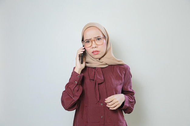 Droevige jonge Aziatische moslimvrouw die een bril draagt en belt met een mobiele telefoon geïsoleerd
