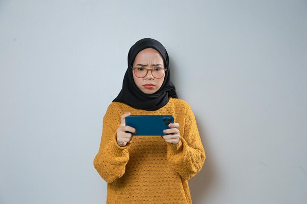 droevige jonge aziatische moslimvrouw die oranje sweater en glazen draagt die spel op mobiele telefoon speelt