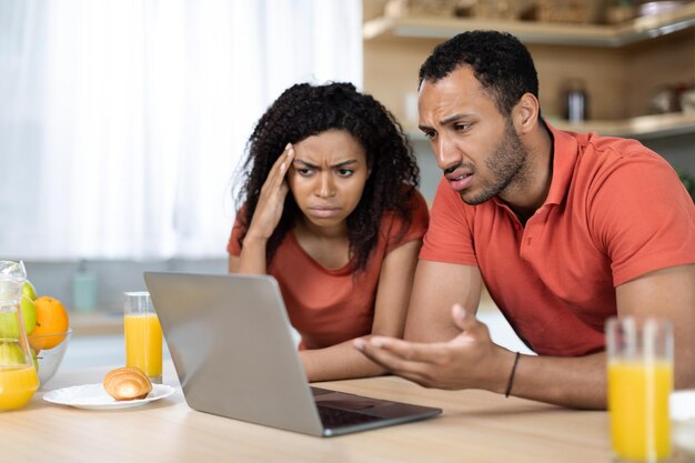 Droevige ongelukkige zwarte vrouw en man in rode t-shirts die naar de computer kijken, hebben problemen met gadgetservice en rekeningen