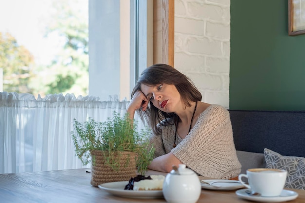 Droevige peinzende of vermoeide vrouw zit aan tafel in café met kopje koffie en dessert Jonge vrouw in gezellig café