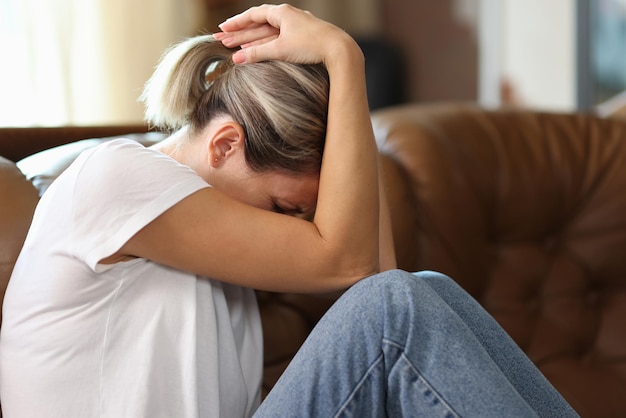 Droevige uitgeputte vrouw die haar hoofd met haar handen vasthoudt terwijl ze thuis op de bank lijdt