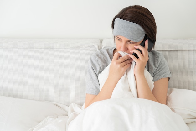 Droevige vrouw die slaapmasker draagt die smartphone gebruikt aangezien zij in bed zit dat met dekbed wordt behandeld