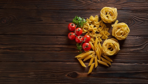 Droge pasta penne, farfalle, tagliatelle met basilicum en cherrytomaatjes op houten tafel. Bovenaanzicht met kopie ruimte