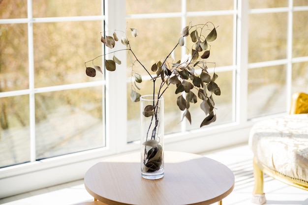 Droge tak met bladeren in een glazen vaas op een witte tafel voor een groot raam.
