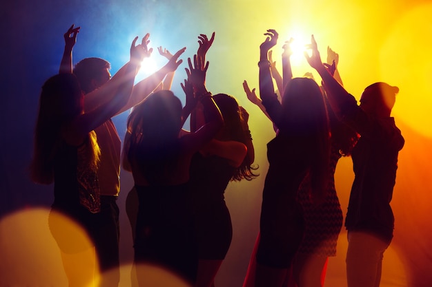 Dromen. Een menigte van mensen in silhouet steekt hun handen op dansvloer op neonlichtachtergrond. Nachtleven, club, muziek, dans, beweging, jeugd. Geelblauwe kleuren en bewegende meisjes en jongens.