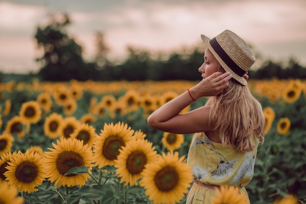 Dromen jonge vrouw in gele jurk met haren en hoed met hand in een veld met zonnebloemen in de zomer, uitzicht vanaf haar kant. Opzij kijken. kopieer ruimte