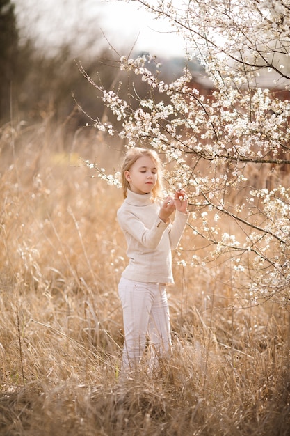 Dromend blondemeisje met gesloten ogen in de bloesemtuin dichtbij de witte bloemen van sakura. lente kersenboom en warme zonnige thema