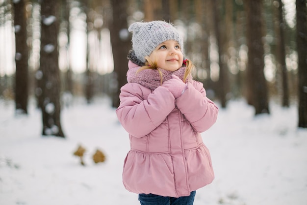 Dromerig schattig klein meisje in stijlvolle warme winterkleren, buiten poseren in het besneeuwde bos en omhoog kijken. Mooi kind meisje in roze outfit genieten van winterseizoen en sneeuw in de natuur.