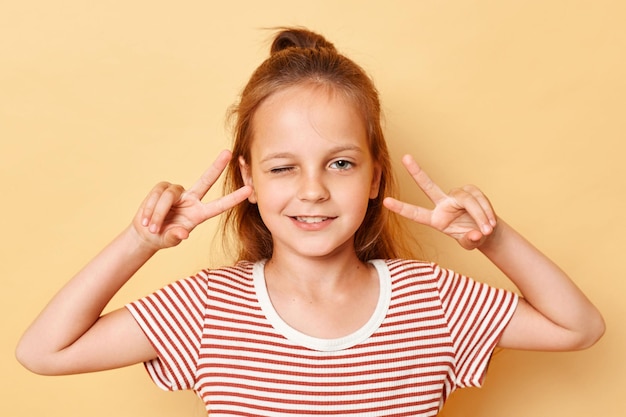 Dromerig vrolijk donkerharig meisje dat vsign toont in de buurt van de ogen knipogend met een casual stijl gestreepte T-shirt die geïsoleerd staat over een beige achtergrond