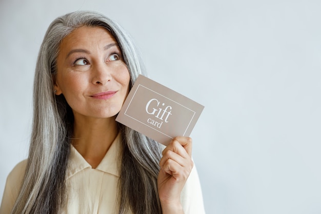 Dromerige grijze haired aziatische vrouw houdt cadeaubon in de buurt van gezicht staande op lichtgrijze achtergrond in studio, ruimte voor tekst. winkelcertificaat