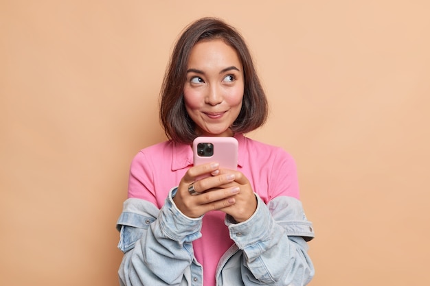 Dromerige jonge mooie Aziatische vrouw met donker haar houdt mobiele telefoon heeft doordachte uitdrukking denkt over ontvangen bericht draagt roze t-shirt spijkerjasje kijkt weg geïsoleerd over beige muur