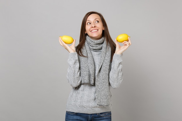 Dromerige jonge vrouw in grijze trui, sjaal opzoeken, met citroenen geïsoleerd op grijze muur achtergrond. Gezonde mode-levensstijl, oprechte emoties van mensen, concept van het koude seizoen. Bespotten kopie ruimte.