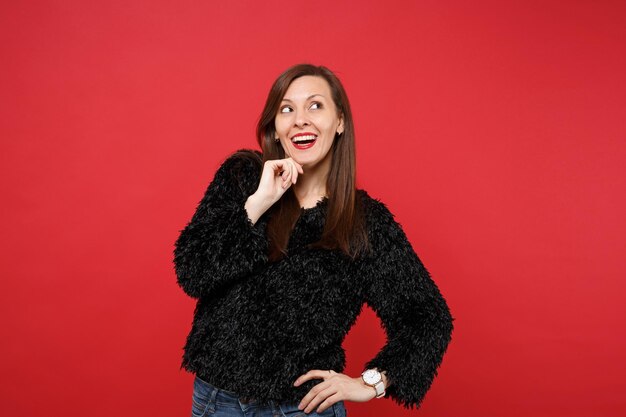 Dromerige jonge vrouw in zwarte vacht trui opzoeken, hand prop op kin geïsoleerd op heldere rode muur achtergrond, studio portret. Mensen oprechte emoties, lifestyle concept. Bespotten kopie ruimte.