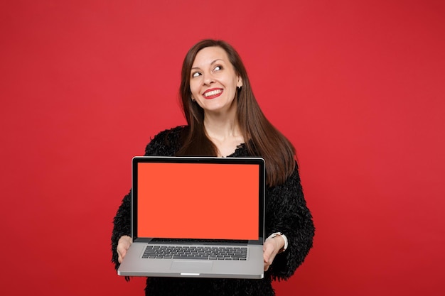 Dromerige jonge vrouw in zwarte vacht trui opzoeken met laptop pc-computer met leeg leeg scherm geïsoleerd op rode muur achtergrond. Mensen oprechte emoties, lifestyle concept. Bespotten kopie ruimte.