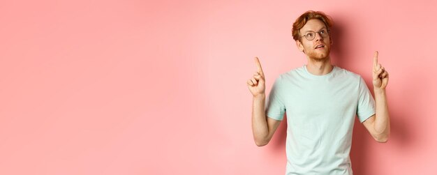 Foto dromerige jongeman met rood haar en baard die met de vingers omhoog wijst en peinzend naar de top kijkt terwijl hij tegen