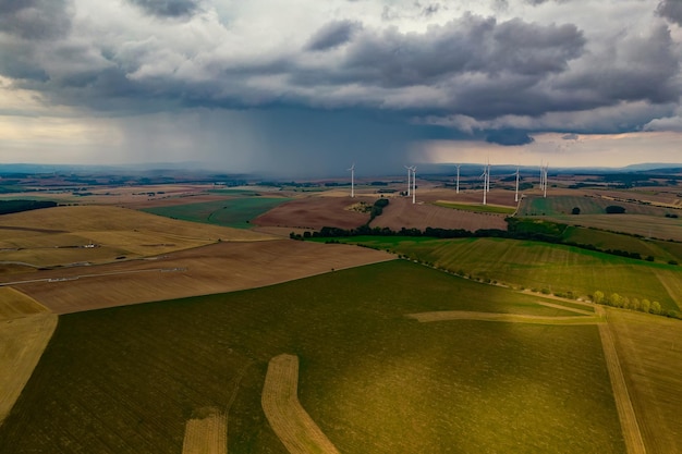 Drone flight at high altitude over a large field with many wind turbines spinning The wind turbines cast a long shadow because the sun is setting In the background at storm sky