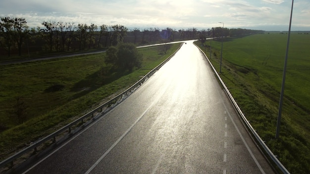 春の朝、播種された畑の間の車のための濡れたアスファルト道路の上のドローン。反射する太陽、太陽のまぶしさのある濡れたアスファルト道路。緑の野原の間の雨の後の濡れた道路。空中ドローンビューフライト