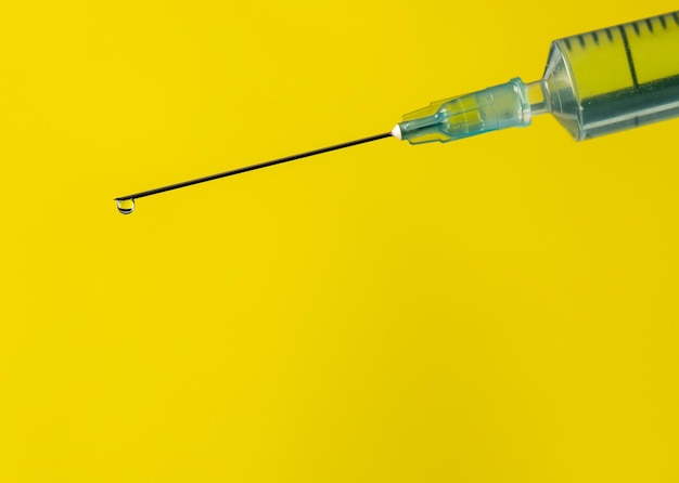 コピースペースのある黄色の背景にある注射器の鋭い針の上に、透明で純粋な水または薬を滴下します。ヘルスケアおよび製薬業界の概念。