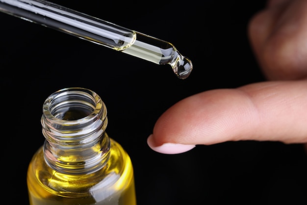 drop of yellow oil flowing from dropper onto woman finger on black background closeup