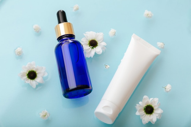 Dropper serum and tube cosmetics products on blue water surface with flowers. Face cosmetics for skin care moisture balance. Aqua cream tube and collagen or hyaluronic serum.