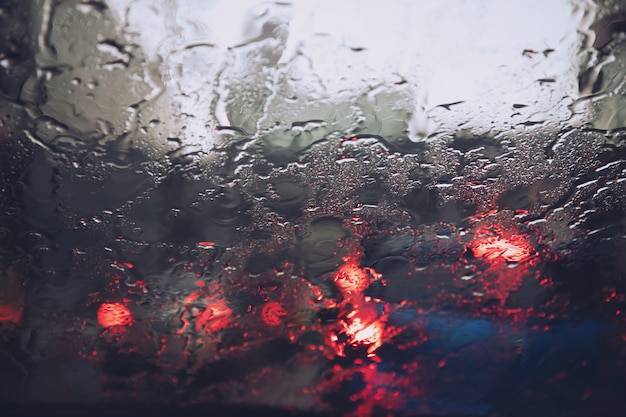 Drops Of Rain Drizzle on the glass windshield in the evening. street in the heavy rain. Tail light and Traffic lights in city.