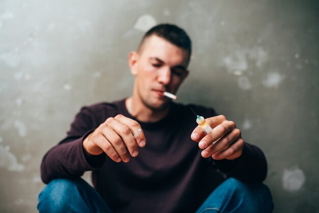 Photo a drug addict with a syringe in his hand and a cigarette in his teeth is sitting on the floor after ...