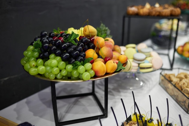 Foto druiven abrikozen ananas mint kersen cakes en macaroons op borden en onderborden op een tafel met een wit tafelkleed tegen een grijze muur in het zuiden tijdens een banket snacks voor het banket