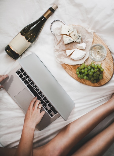 Druiven en een fles witte wijn met kaasplateau voor de lunch in de slaapkamer. De jonge vrouw werkt thuis, gebruikend notitieboekje