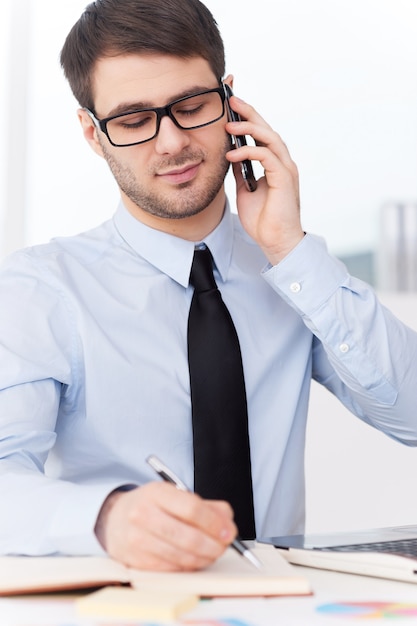 Druk aan het werk. Knappe jonge man in overhemd en stropdas die op de mobiele telefoon praat en iets in notitieblok schrijft terwijl hij op zijn werkplek zit