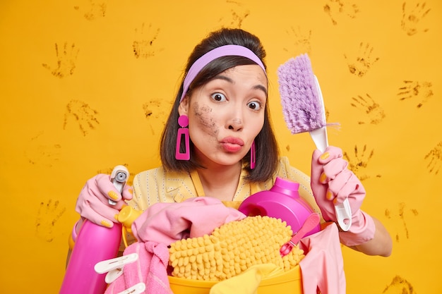 Drukke huisvrouw probeert alles netjes te houden heeft lippen gevouwen die verantwoordelijk zijn voor het doen van dagelijkse klusjes houdt borstel voor afstoffen en schoonmaakmiddel poses in de buurt van wasmand geïsoleerd op gele muur