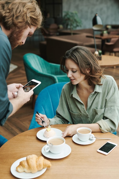 Drukke jonge, bebaarde man zit aan tafel in een modern café en drinkt koffie tijdens het bespreken van het project