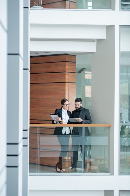 Drukke jonge kantoorpersoneel in formalwear staande op balkon in kantoor centrum en met behulp van tablet tijdens het bespreken van businessplan