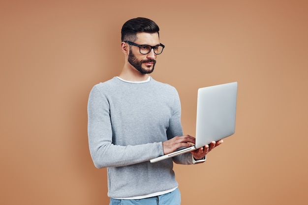 Drukke jonge man in slimme vrijetijdskleding met laptop terwijl hij tegen een bruine muur staat