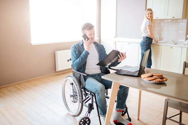 Drukke jonge student op rolstoel studeren en telefoneren. Man met speciale behoeften en een handicap. Geopende map vasthouden. Jonge vrouw staan op fornuis en koken