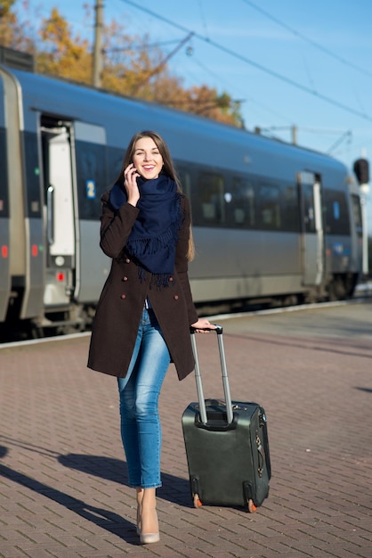 Drukke jonge vrouw in jas met koffer praten aan de telefoon op het treinstation