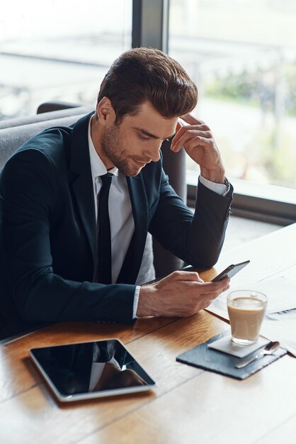 Drukke jonge zakenman in volledig pak die smartphone gebruikt terwijl hij in het restaurant zit