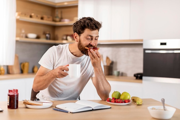 Drukke knappe volwassen blanke man in wit t-shirt eet sandwich met jam drinkt koffie leest boek