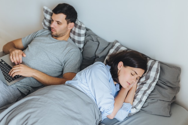 Drukke man ligt in bed, werkt op laptopcomputer, heeft ernstige uitdrukking terwijl zijn vrouw een gezonde slaap heeft