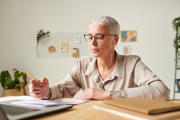 Drukke mooie zakenvrouw in oortelefoons die aan het bureau op kantoor zitten en informatie in kranten onderzoeken