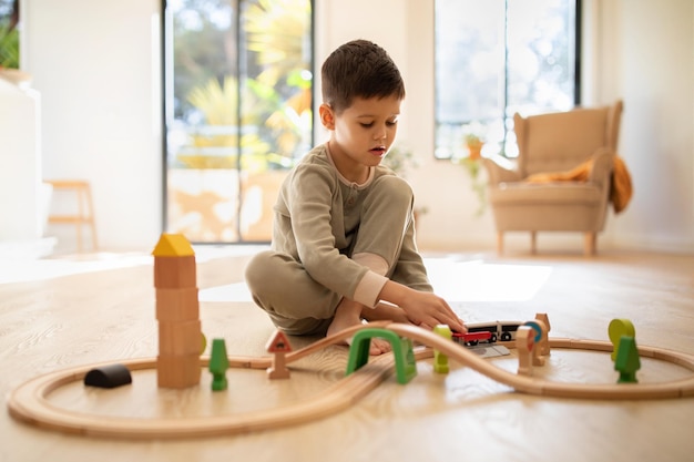Foto drukke, serieuze blanke kleine jongen bouwt stad, speelt houten wegtrein en auto's in het interieur van de kinderkamer