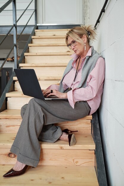 Drukke volwassen vrouw in stijlvolle outfit zittend op de stap van houten trap en analyseren van gegevens op laptop