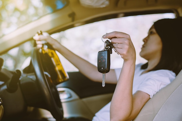 도로에서 차를 운전하는 동안 차 열쇠를 들고 술을 마시는 취한 아시아 여성여성들은 맥주 한 병으로 차를 운전한다위험한 운전 개념